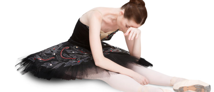 Ballerina in a black-and-red tutu, tights and pointe shoes sits on the floor resting her head on her hand. She looks exhausted and upset.