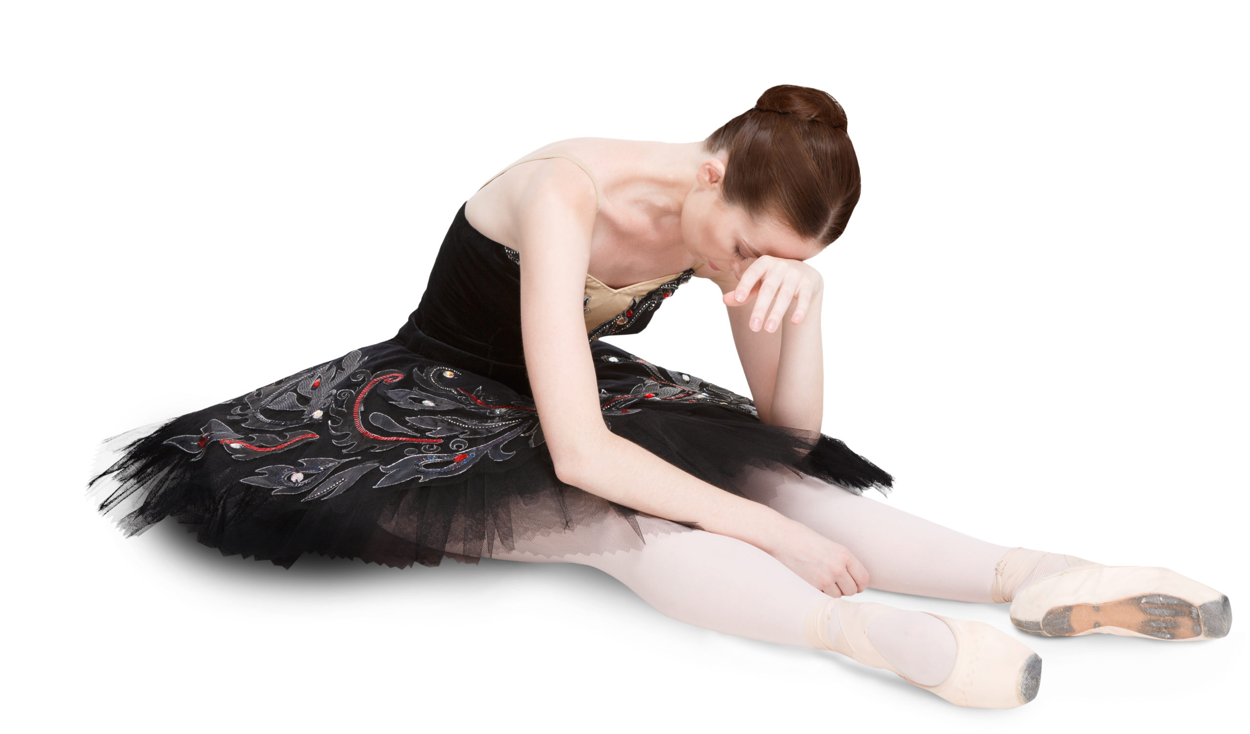 Ballerina in a black-and-red tutu, tights and pointe shoes sits on the floor resting her head on her hand. She looks exhausted and upset.