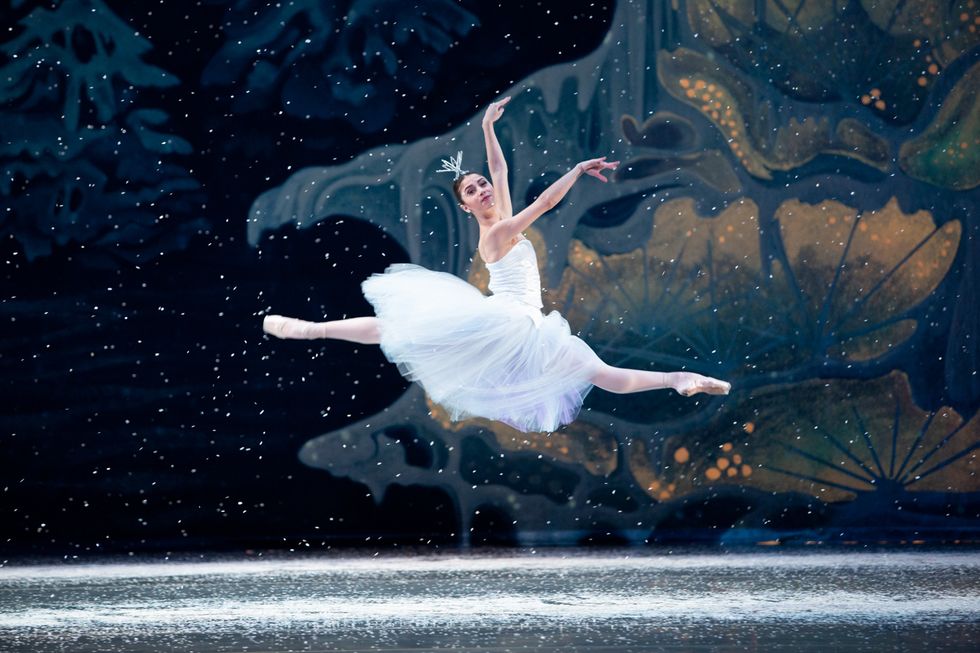 Wearing a long white tutu and crown, Nina Fernandes does a saut de chat in front of a wintery backdrop as snow falls from the top of the stage.