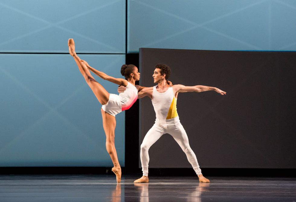 A male dancer catches a female dancer in his right arm as she wraps her left arm around his shoulder and executes a high arabesque on pointe. Both wear white costumes and dance in front of a blue backdrop onstage.