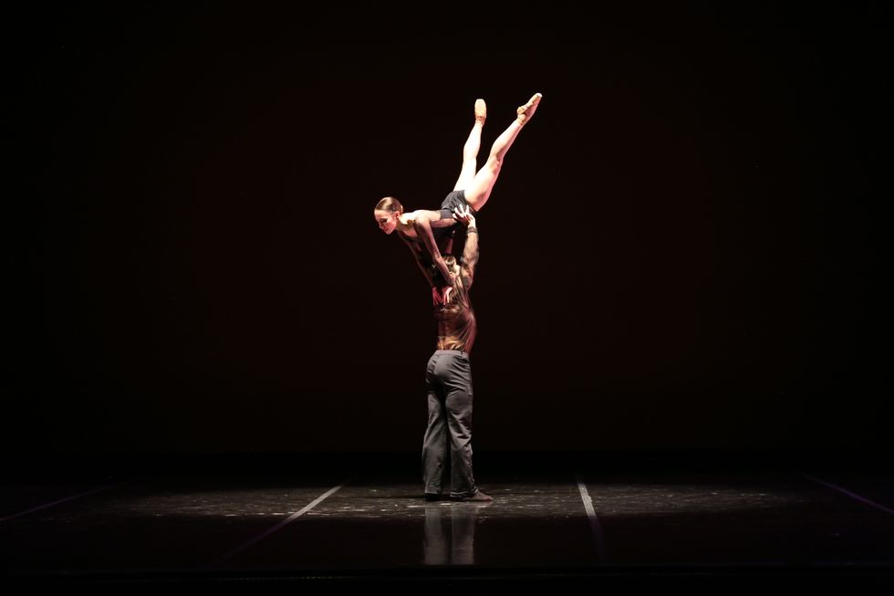 A shirtless male dancer in gray pants lifts a female ballerina above his head as she holds onto his shoulders and extends her legs straight into the air.
