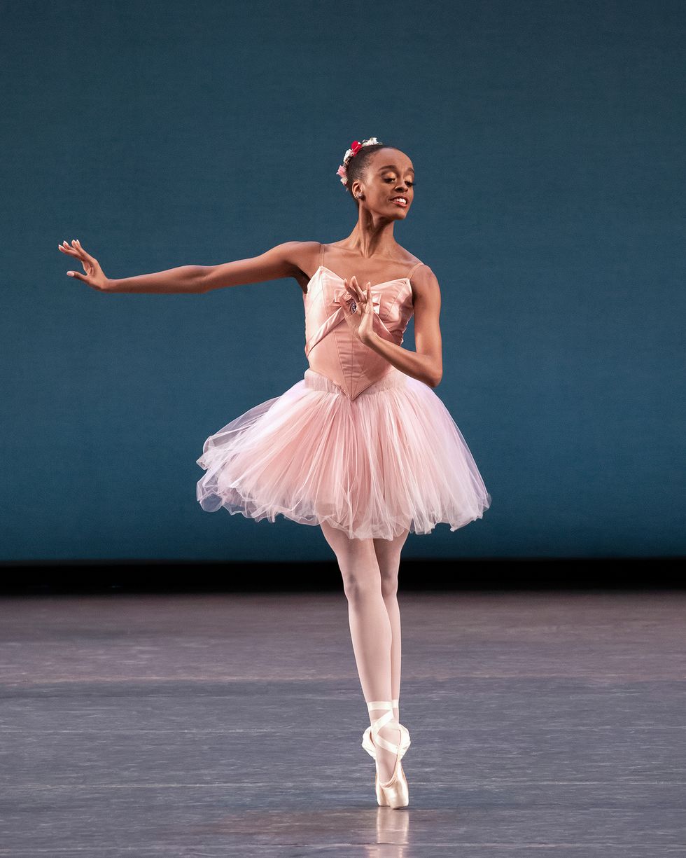 India Bradley, dressed in a pink tutu, tights and pointe shoes and wearing a flowered wreath around her bun, poses in sous-sus with her left hand flexed in front of her chest.