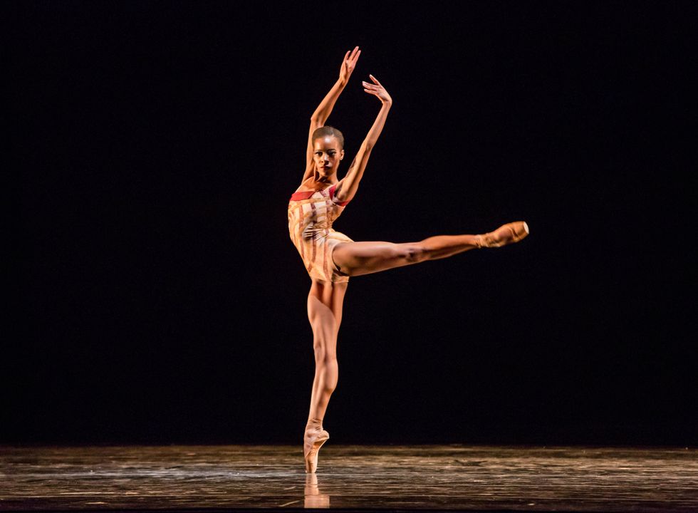 Wearing a beige and brown striped leotard with red embellishments, Dara Holmes does relevu00e9 arabesque on her right leg and lifts both arms high into fifth position.