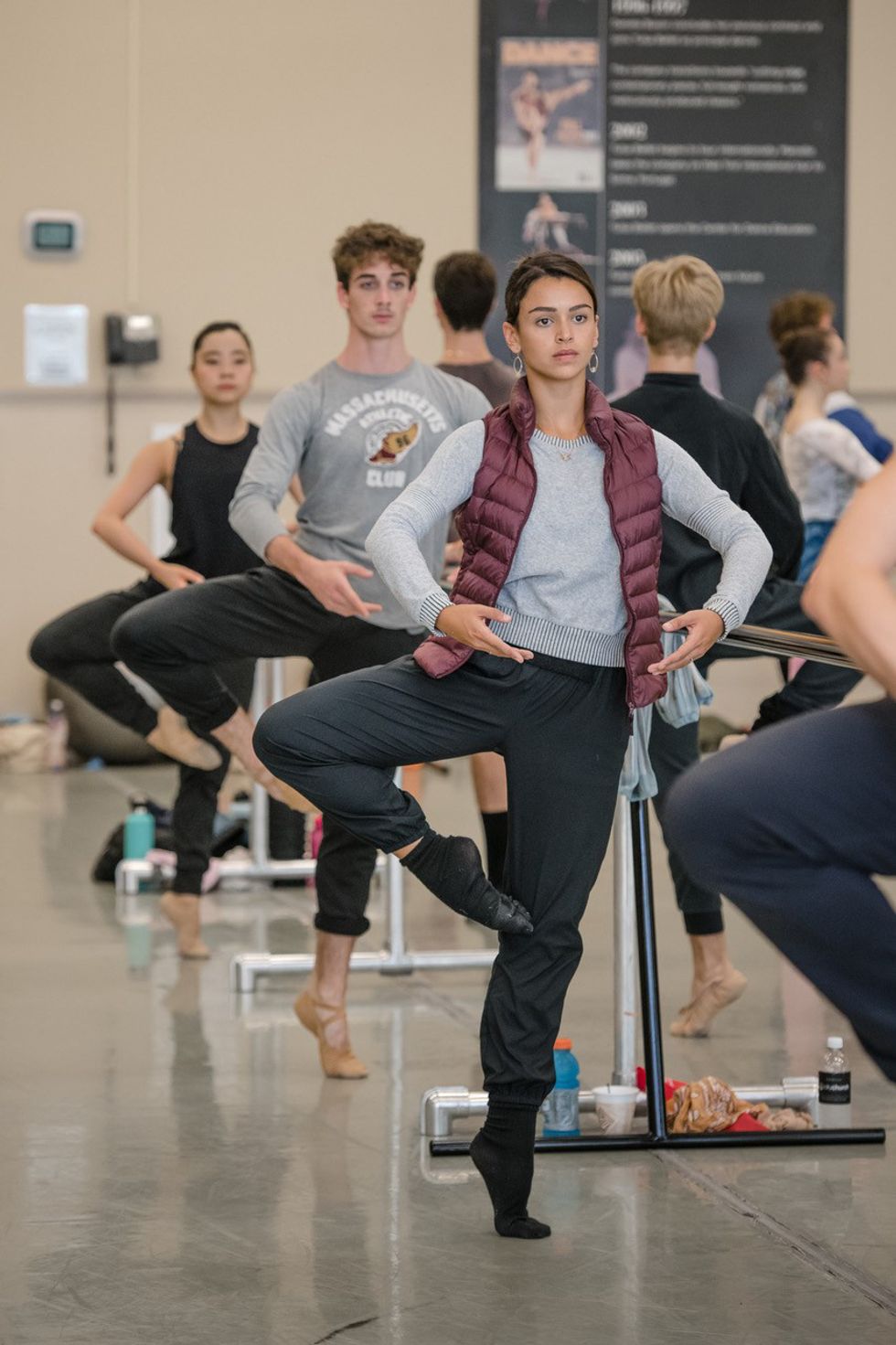 Wearing black exercise pants, a gray sweatshirt and burgundy puffy vest, A female ballet dancer balances in passu00e9 at the barre during a ballet class.