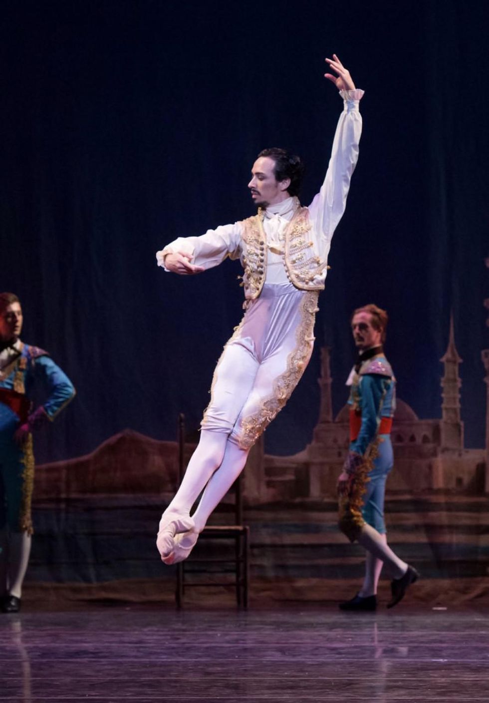A white male danseur in a white and gold Spanish-style bolero jacket and tights, is shown onstage doing an assemblu00e9 to the right.