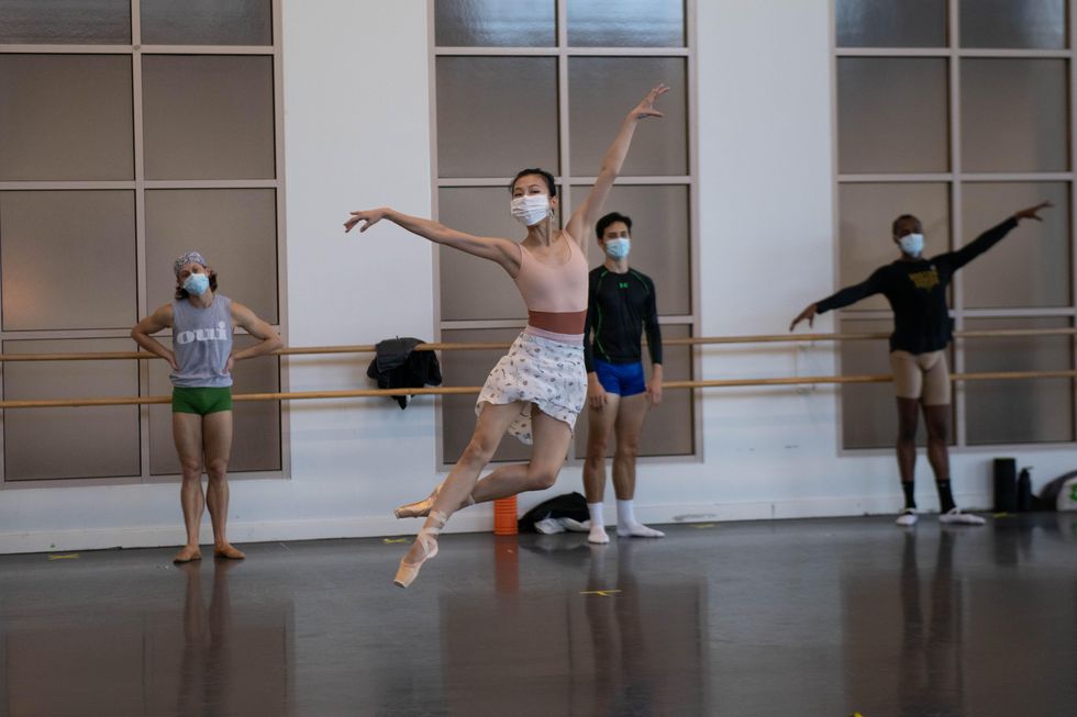 An Asian ballerina in a face mask, pink leotard, white skirt and pointe shoes does a jetu00e9 sautu00e9 with her left foot back in coupu00e9. Behind her three male dancers in practice clothes wait near the barre.