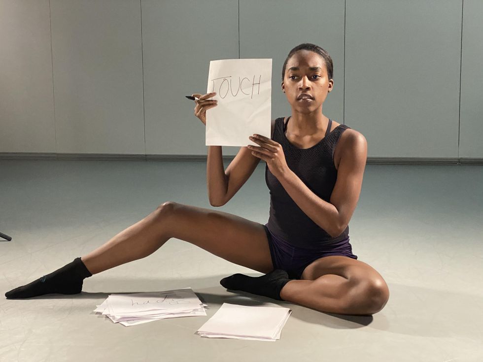 A young Black ballerina in a black leotard, booty shorts and black socks sits on the floor with her left leg tucked in and her right leg extended and bent. She holds up a piece of paper that says "TOUCH."