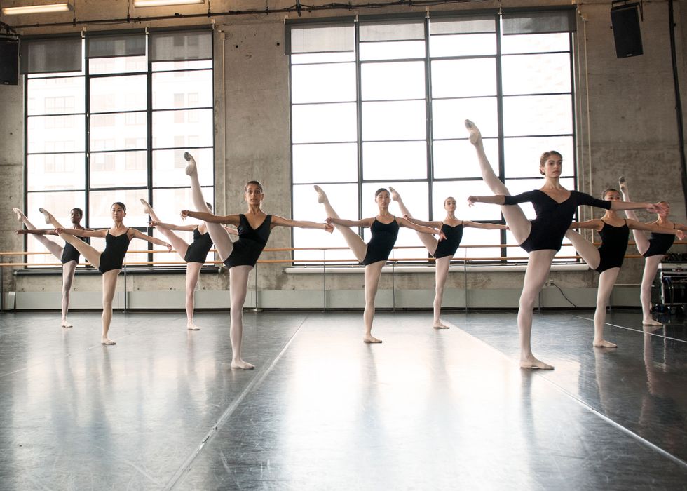 A class of young women in a brightly lit studio with large windows. They have one leg in a la seconde, and their arms in second.