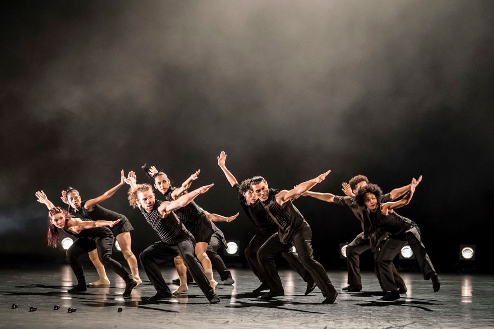 Nine male and female dancers, all dressed in black, move through a deep second pliu00e9, arms flung wide.