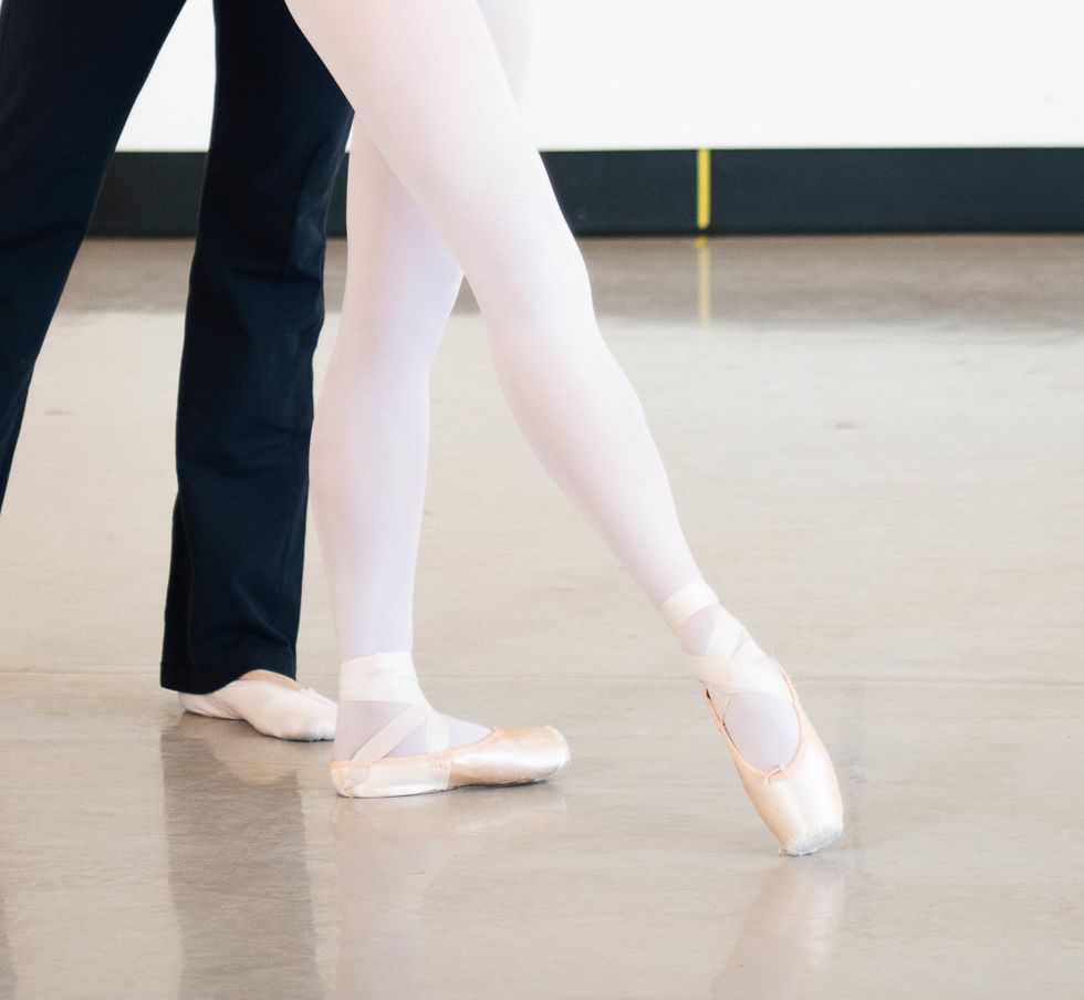 A young female dancer is shown in tendu croisu00e9 devant from the knee down, wearing pink tights and pointe shoes.