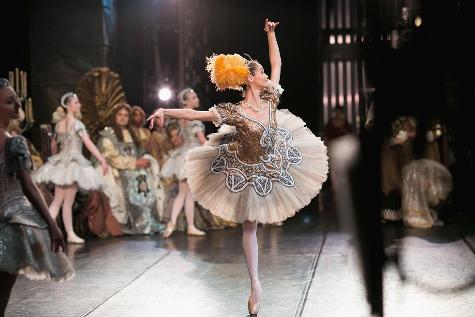 In this photo shot from offstage right, a ballerina in an orange-tone tutu and feather plume headpiece does a piqué arabesque on her right leg and looks out towards the audience, smiling.
