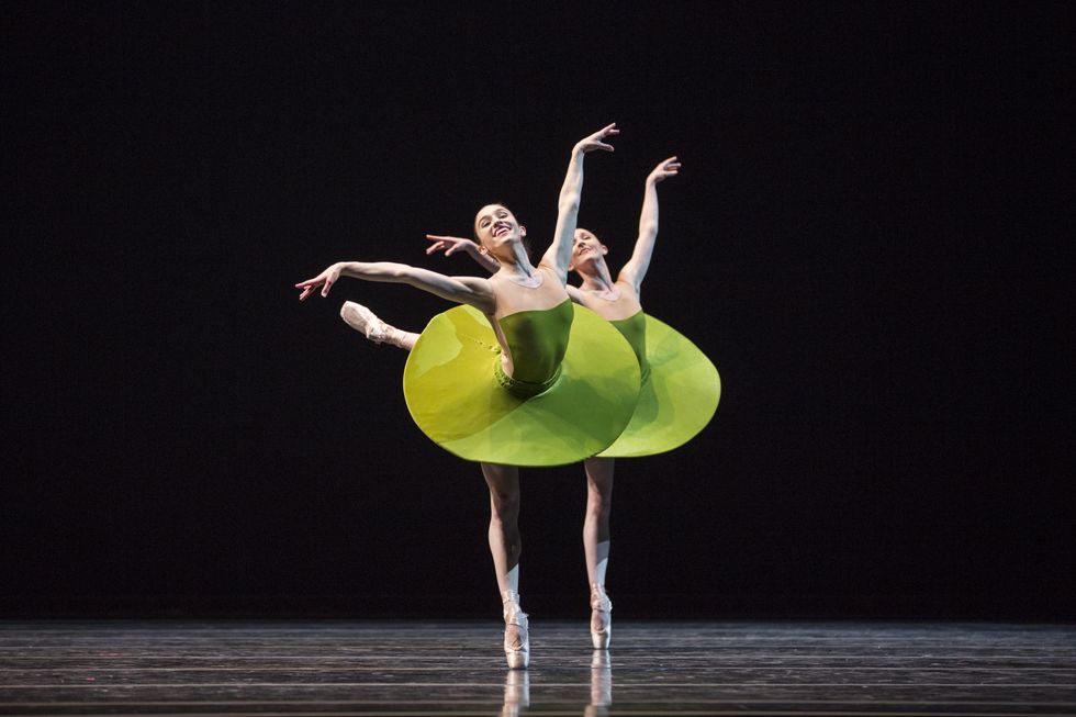 Two ballerinas, one in front of the other, piqué arabesque croisé on their right leg with their left arm raised high and their right arm out to the stand. They dance on a darkened stage and wear lime green disc-shaped tutus.