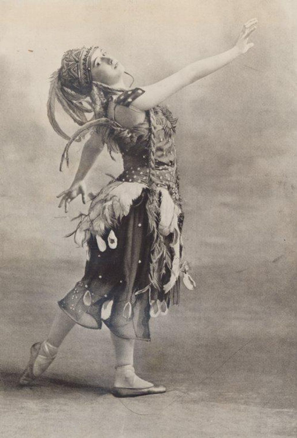 In this antique black-and-white photograph, a ballerina wears a tall feathered hat, an ornate bodice and loose, calf-length pants covered in feathers and sparkly adornments. She stands in profile, in a slightly turned in B+ position and stretches her right arm out in front of her.