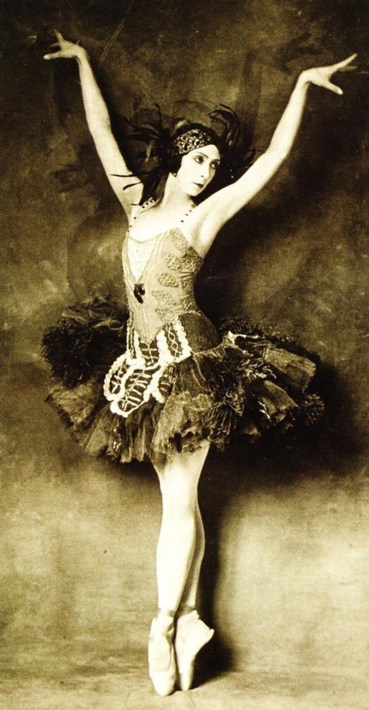 In this antique, sepia-toned photograph, a ballerina stands in sous-sus on pointe with her arms raised in a V position and looks towards her left. She wears a dark, dropped waist tutu covered in lace and feathers.