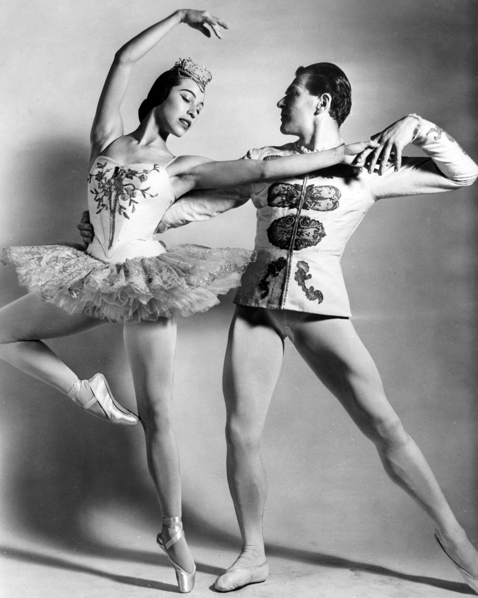 In this black-and-white photo from 1957, a ballerina in a very short, light-colored tutu and tiara stands in retiré and pulls off her left leg slightly. Her partner, in white tights and princely tunic, holds onto her waist and left hand and stretches his left leg out in tendu.