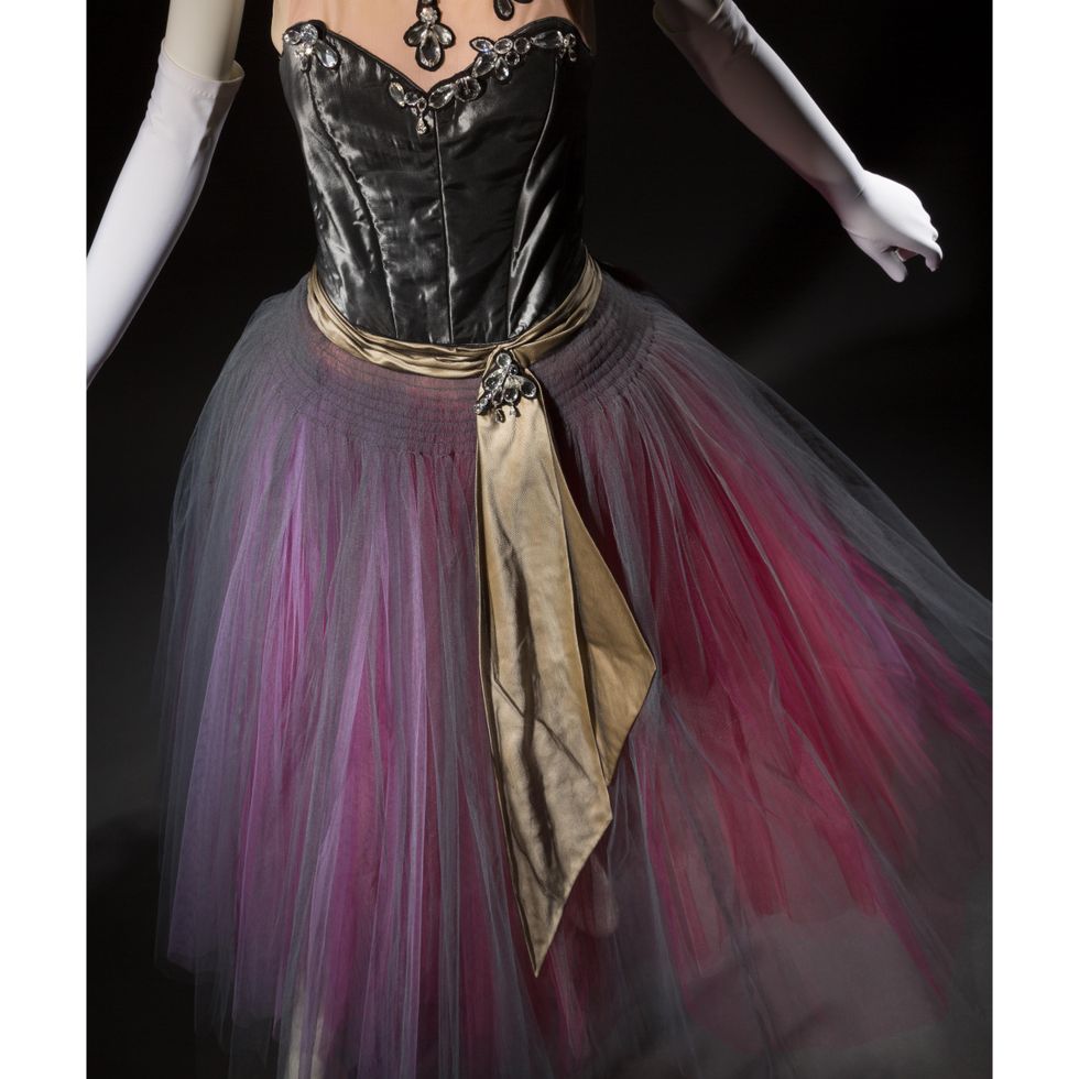 This close up photo shows a long Romantic-style tutu with a gray velvet bodice, gold sash, and skirt made from layers of gray, pink and red tulle.