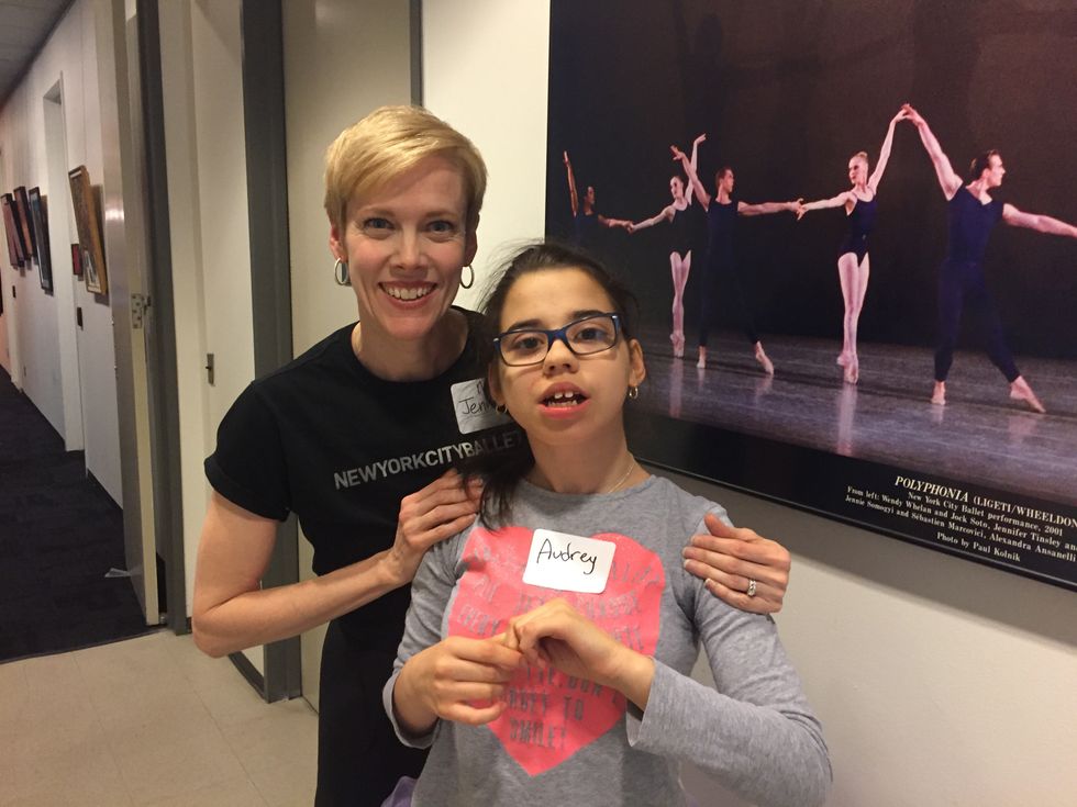 A woman with short blond hair wearing a New York City Ballet shirt poses behind Audrey, who wears glasses and a name tag.