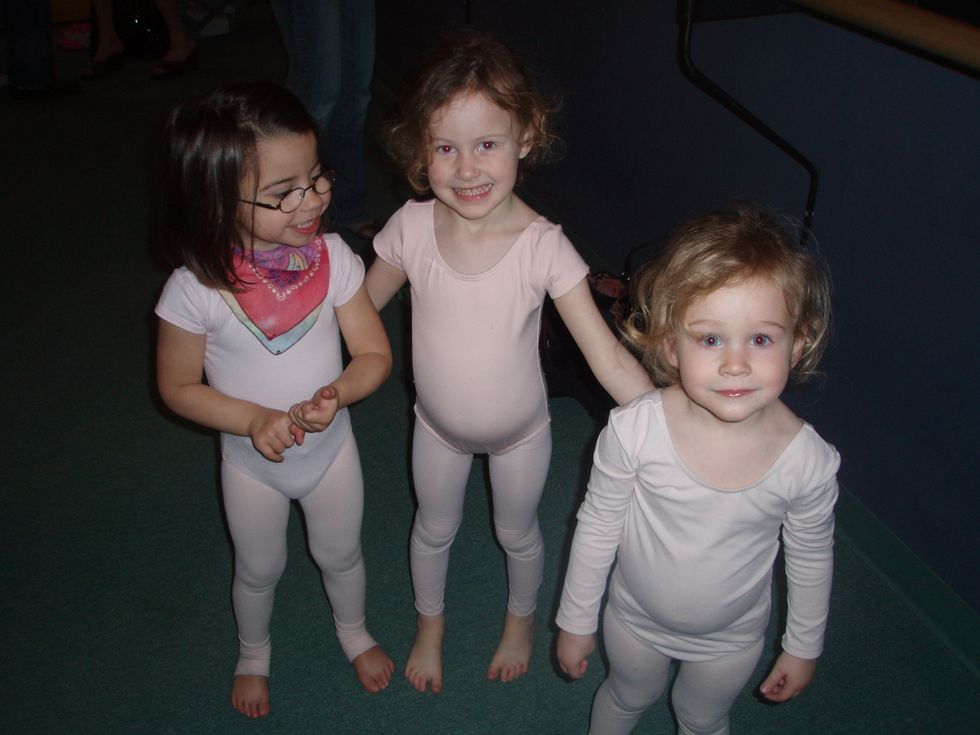 Three four-year old dancers in pink leotards and tights stand in a row and smile at the camera. Audrey wears glasses and a bandana around her neck.