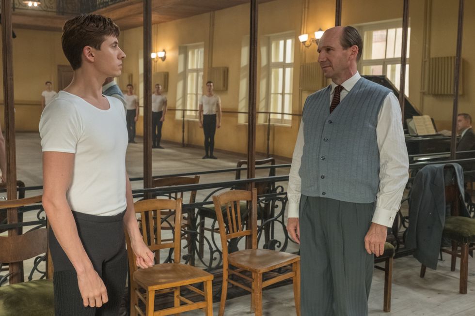 In a scene from The White Crow, Rudolf Nureyev stands at attention in a ballet uniform as his teacher, Alexander Pushkin, considers him. Behind them is a mirror, in which other male ballet students are standing at attention at the barre and an accompanist awaits instructions at his piano.
