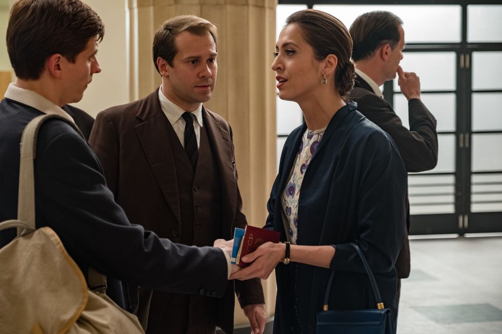 In a scene from The White Crow, a woman grasps Rudolf Nureyev's hand in both of hers, their expressions conflicted. A suited KGB agent stands between them.