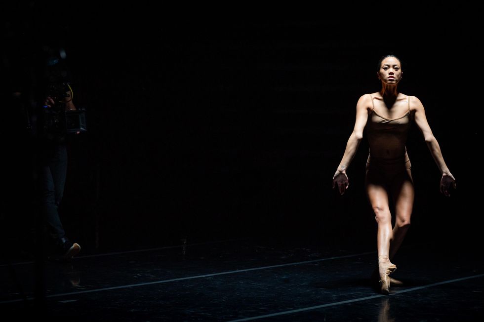 Jeraldine Mendoza, wearing a brown bra-top and briefs and pointe shoes, stands in tendu devant en face on her left leg. Her arms reach straight down with flexed hands as light from above pools around her on the darkened stage.