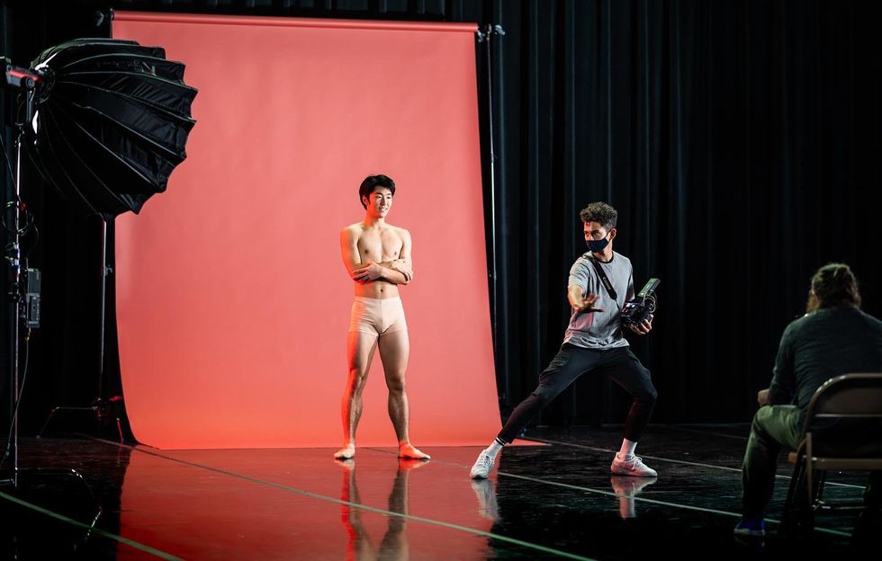 Hyuma Kiyosawa wears nude booty shorts and stands in front of a pink backdrop with his arms crossed at his bare chest. He watches Xavier Nu00fau00f1ez, who is in front of him in street clothes and lunges ardently onto his left leg. He has a camera looped around his chest and looks over his right shoulder towards Kiyosawa.
