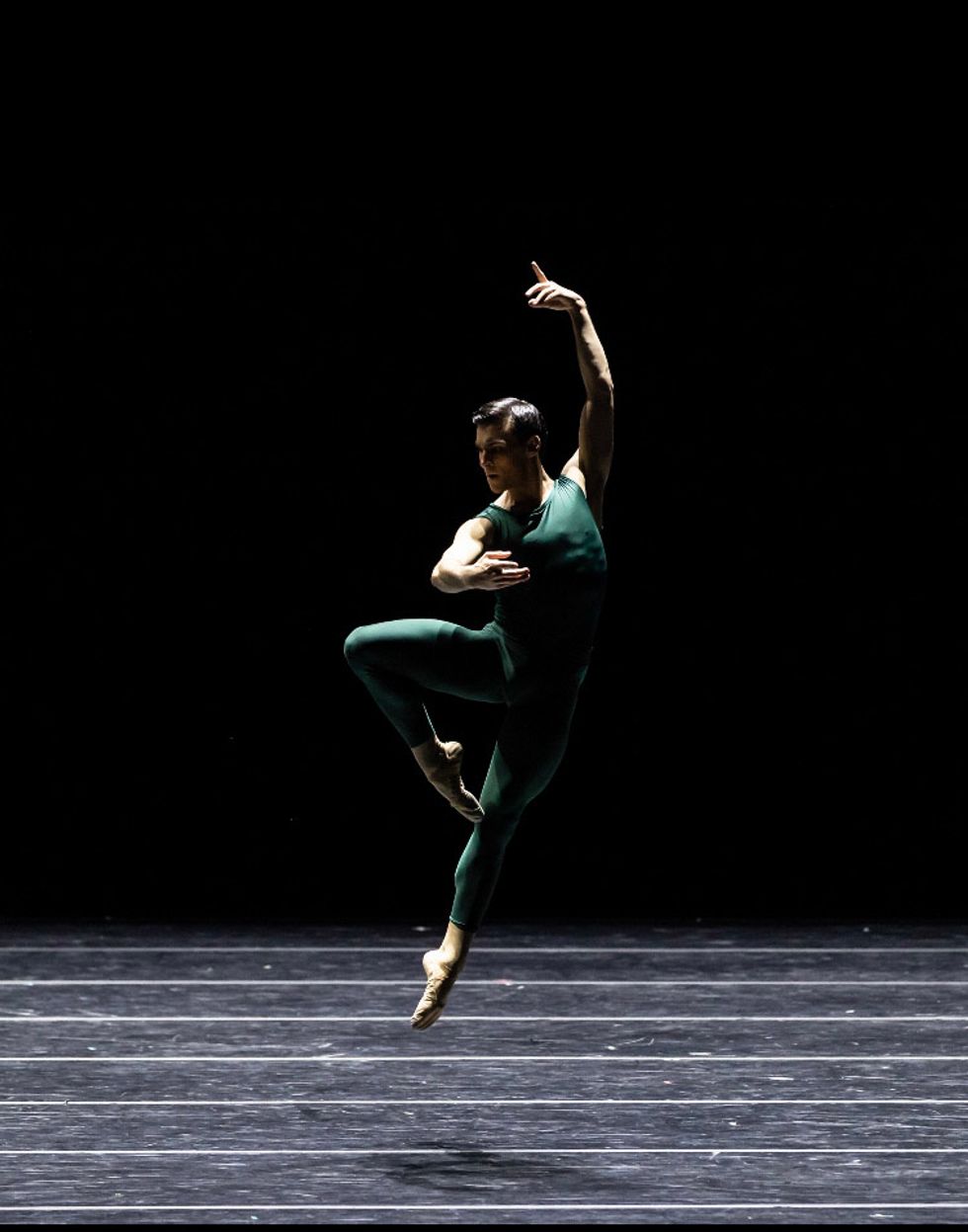 Wearing a green unitard, Cameron Thomas performs a sautu00e9 jump with his right leg in retiru00e9 and his left arm held above his head.