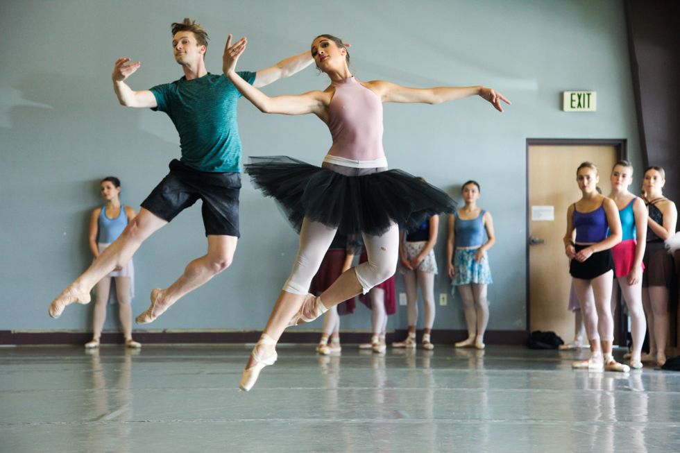 Two dancers in rehearsal clothes jump in jetu00e9s in class with other dancers standing behind them