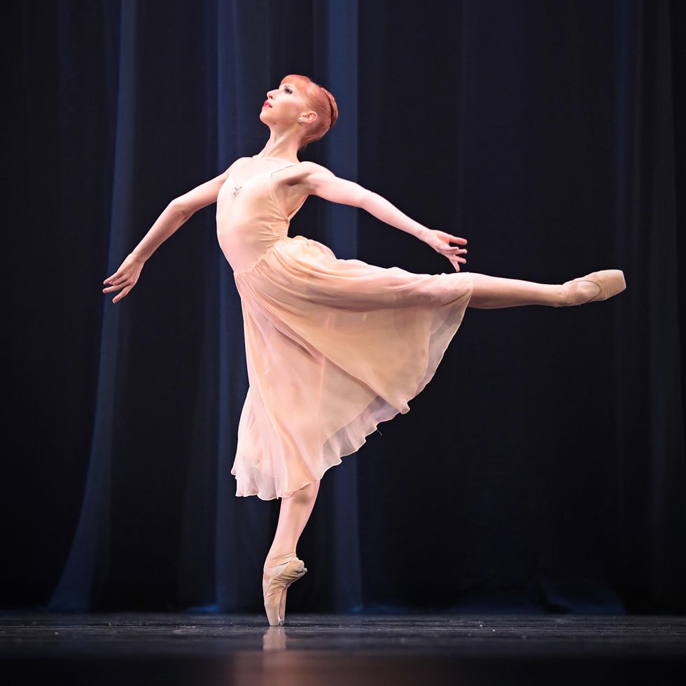 A young ballet dancer in a long pink dance dress and pink tights poses in arabesque on pointe on her right leg in front of a dark curtain. Both arms are held out to the side, with her right arm curling under counter-clockwise and her left arm curling up