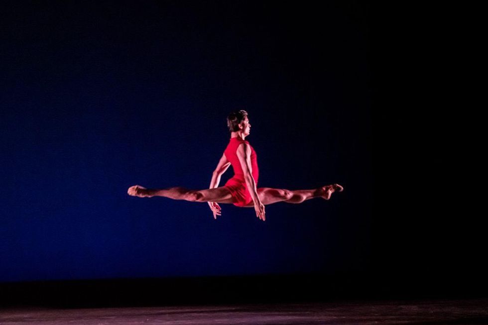 A young male ballet student in a red biketard is shown leaping across a stage, holding both of his arms down by his sides.