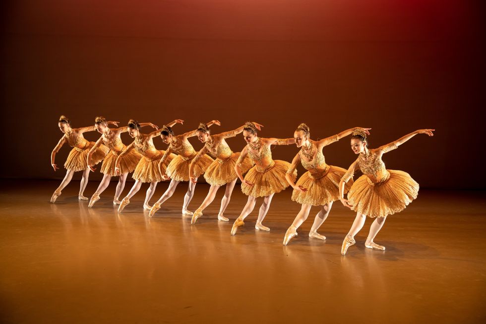 A group of young ballerinas wearing yellow tutus, pink tights and pointe shoes stand in a diagonal line in tendu devant effacu00e9 with their left legs in pliu00e9. Their right arm is curved and their left arm is extended out to the side.