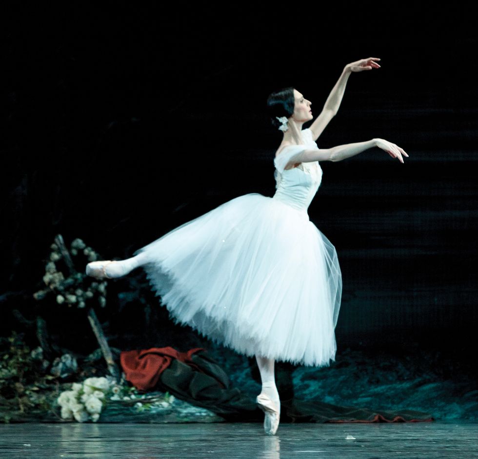 Hodgkinson, in a white tutu, in an arabesque on pointe with her arms in fourth position.