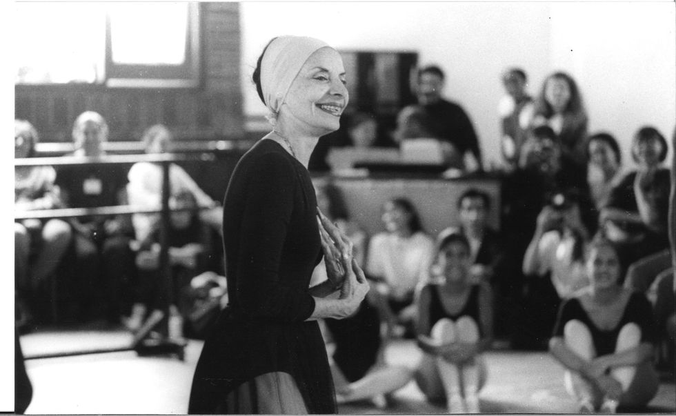 Alicia Alonso, in a long=sleeved black leotard, dark tulle skirt, and headband smiles at out of focus dancers and spectators seated around her as she stands in the middle of a ballet studio.