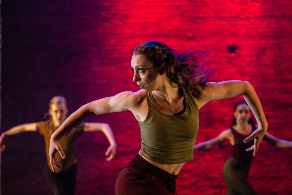 Zou00e9 Emilie Henrot looks sharply to the right and holds her arms out to the side with her elbows bent while twisting her lower body left at the hips. She wears an olive green tank top and burgundy tights, and is flanked by two female dancers behind her on either side doing the same pose. A red light glows on the brick wall behind them.