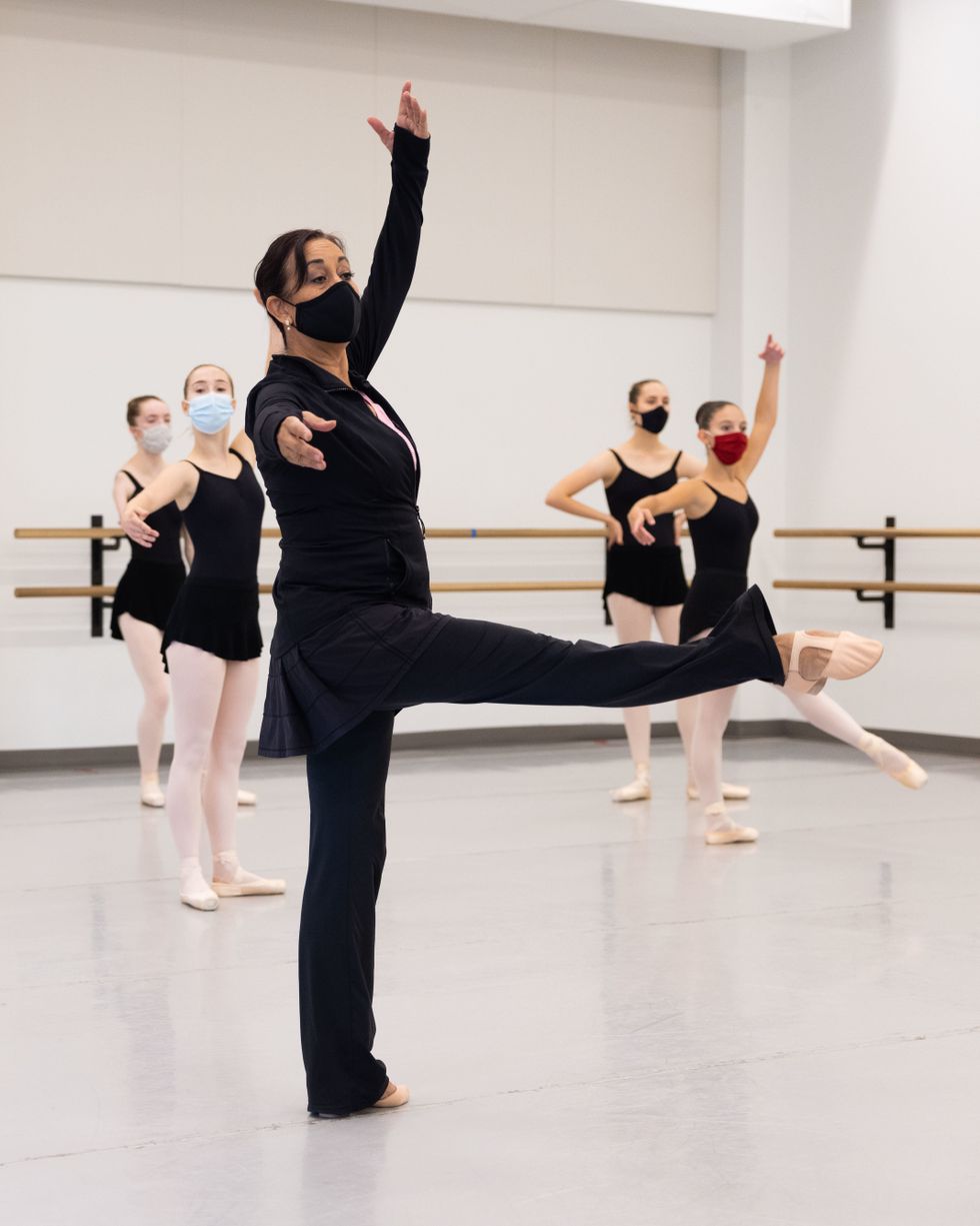 Evelyn Cisneros-Legate extends a leg croisu00e9 front at 90 degrees in center, upstage arm in fifth. Behind her, masked students in pink tights and black leotards watch or imitate. All wear face masks.