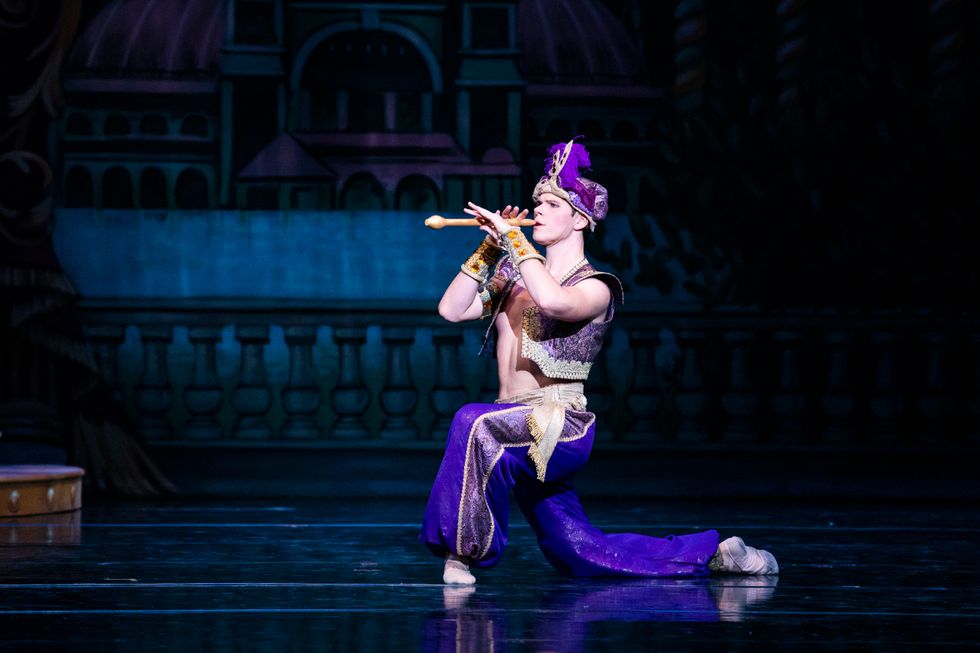 Naumann kneels onstage in purple pants and a vest and headpiece, playing a wooden flute.