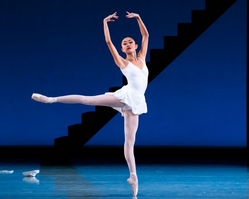 Ohtaki in a white leotard and skirt in an arabesque on pointe onstage with her arms in fifth position.