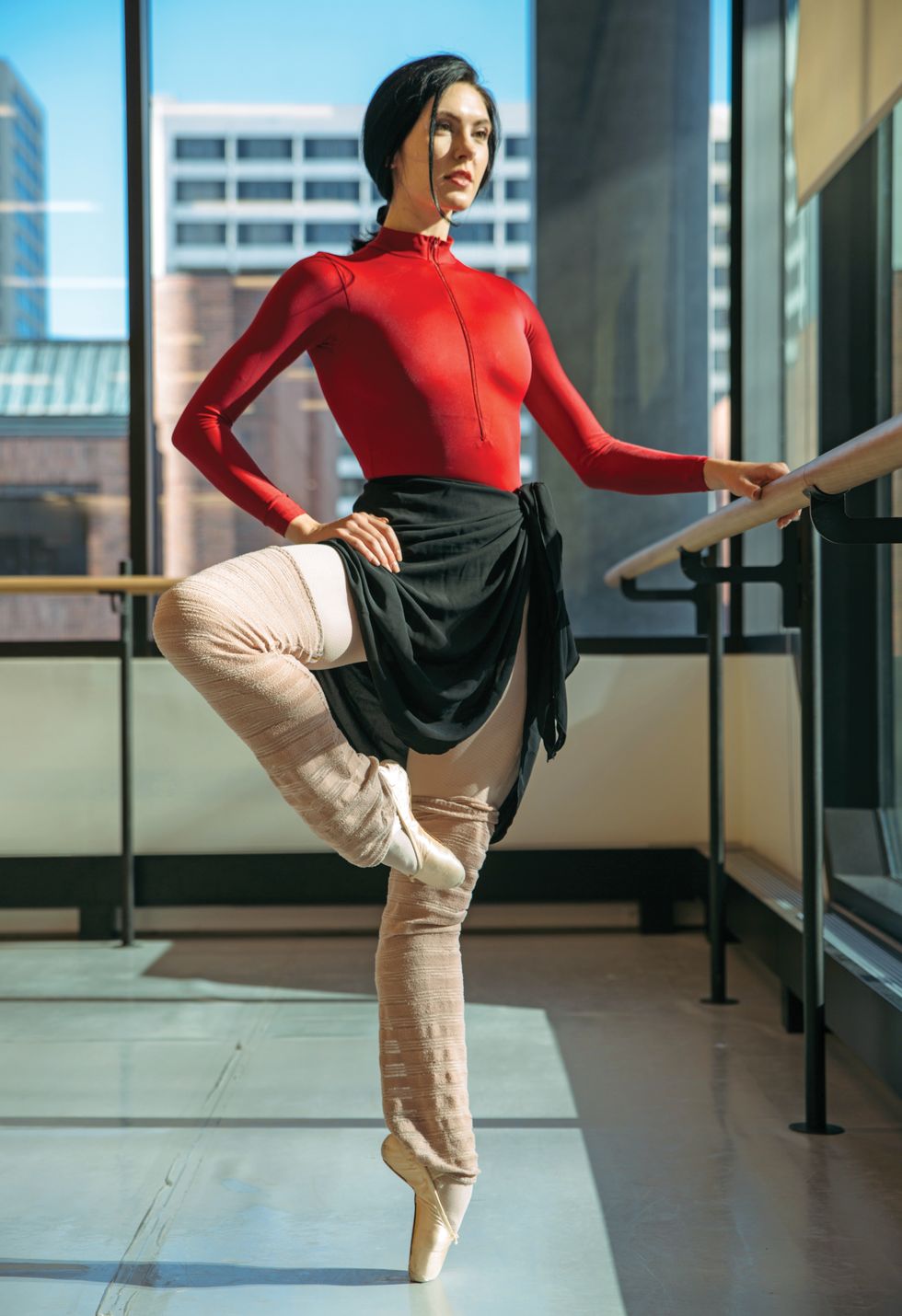 Sisk, dressed in a red leotard and black skirt and legwarmers, stands in a passu00e9 on pointe with one hand on the barre.
