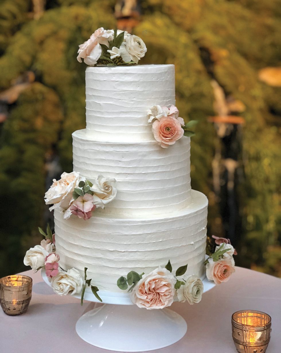 A three-tiered white wedding cake, decorated with pale roses, on a white cake stand on a table with trees in the background.