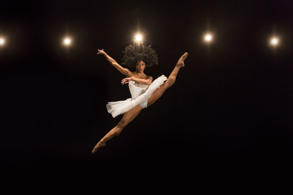 Rachael Parini, wearing a white tutu, does a large split sissone with her legs in second, her left leg higher than her right to create a diagonal. She crosses her left arm in front of her torso and lifts her right arm up to the side past her shoulder.