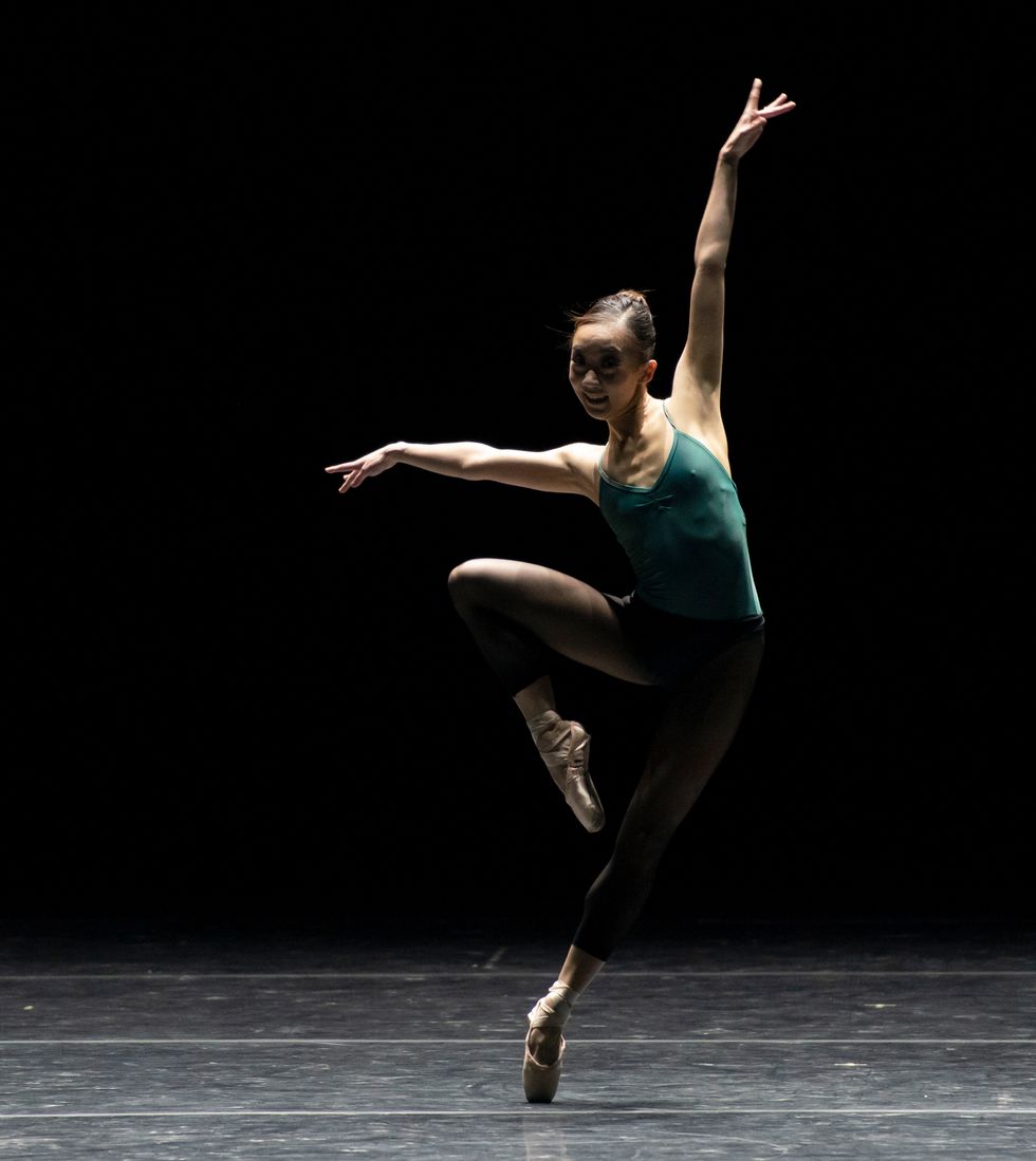 Oga, in a green leotard and black tights and pointe shoes, is onstage in an off-kilter passu00e9 with her arms in a jazzy extension.