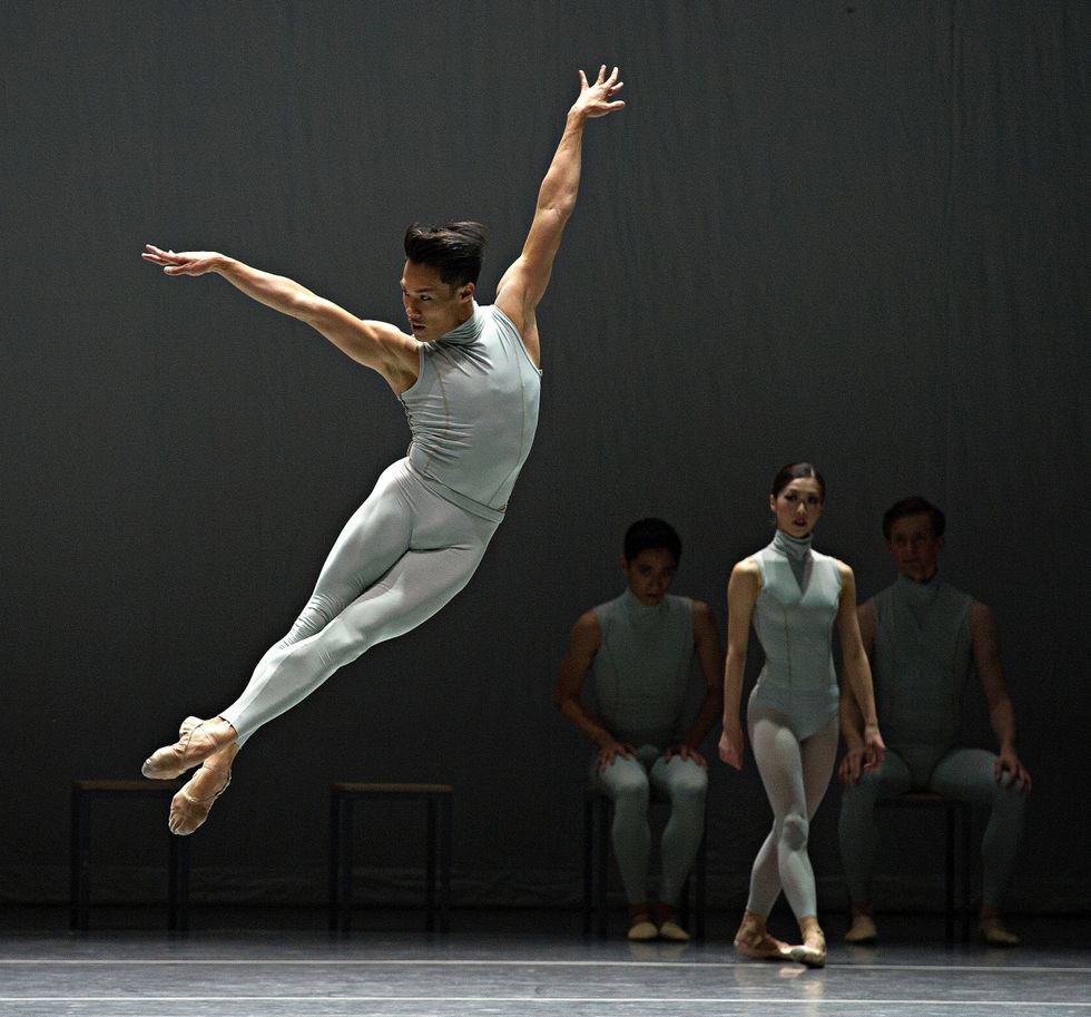Lam in a light grey top and tights leaps in the air onstage with his ankles crossed and his body curved to the side.