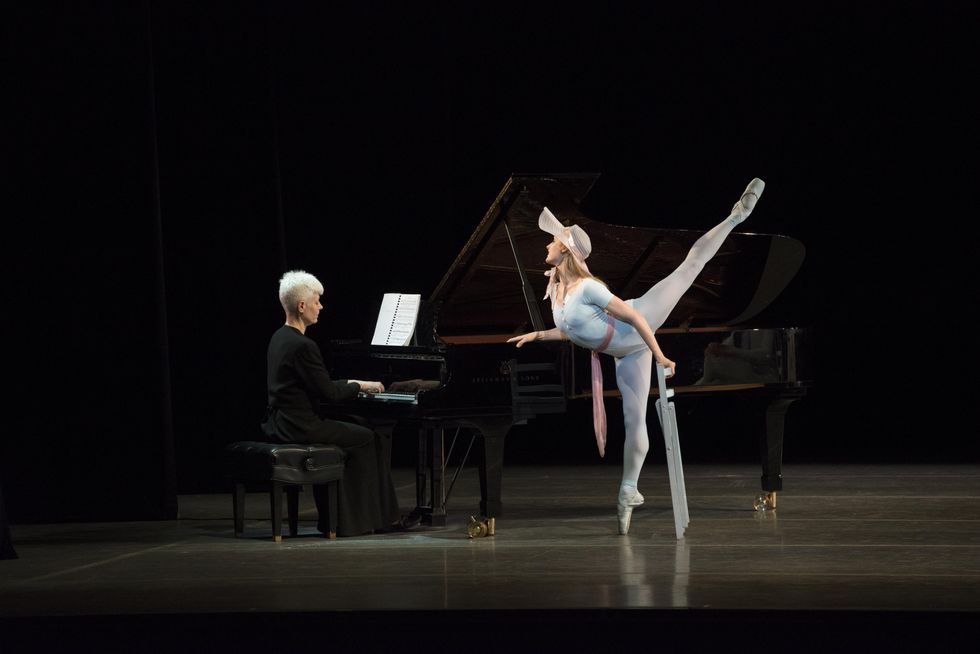 Ashley Ellis, dressed in a floppy hat and a blue leotard, blue tights and pointe shoes, presses down on a black grand piano onstage with her right hand and lifts her left leg high in arabesque allongu00e9 while holding a folding chair in her left hand. She looks attentively toward the pianist, a woman with short white hair dressed all in black..