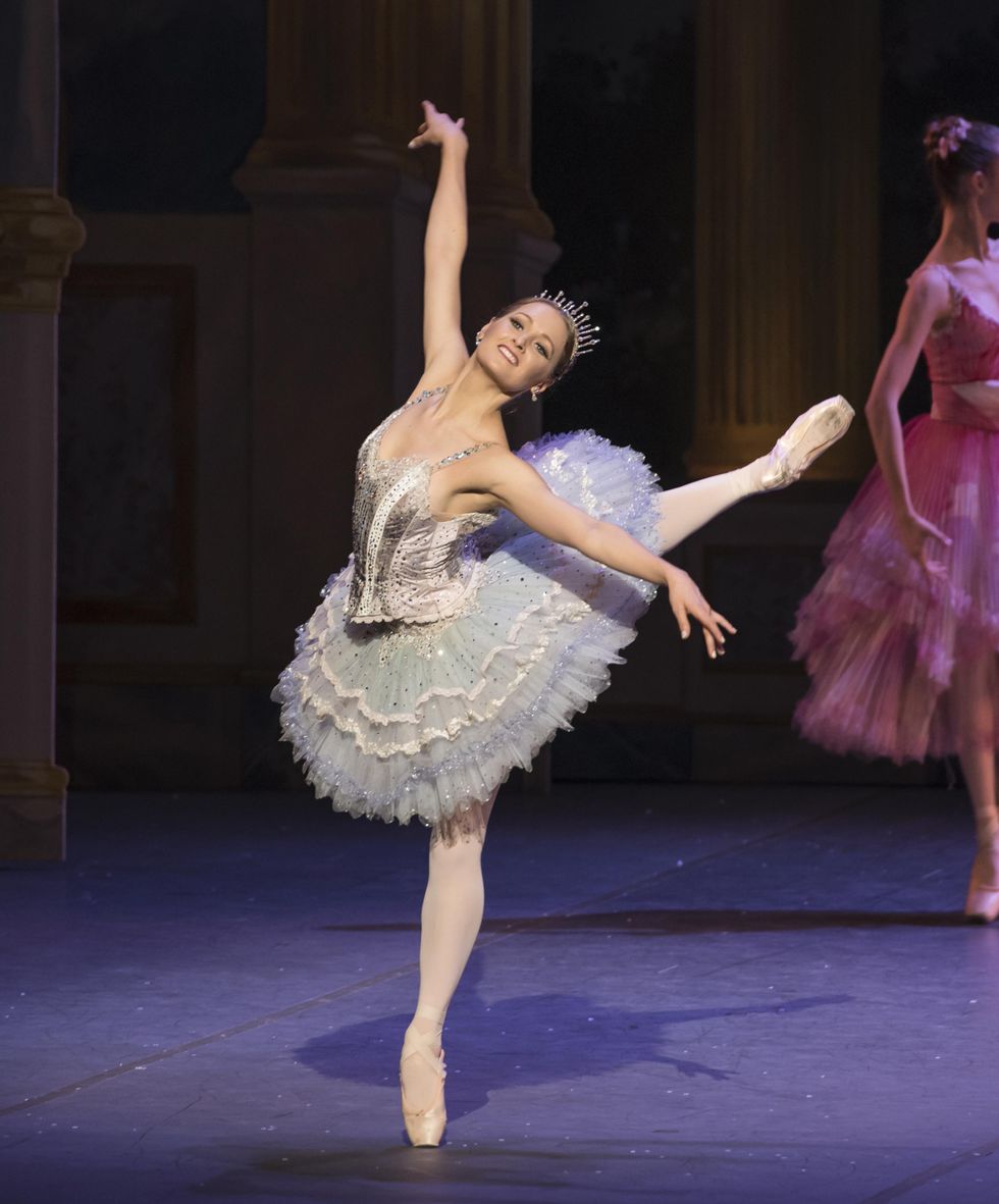 Wearing a sparkly pancake tutu and tiara, Ashley Ellis does a piquu00e9 attitude derriere croisu00e9 on her left leg, lifting her right arm up and her left arm to the side. She arches her upper body out towards the audience and smiles brightly,.