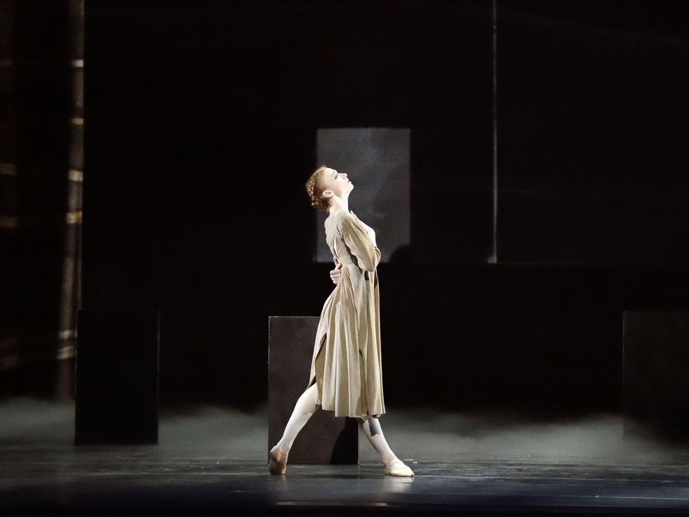 Catherine Hurlin, in a drab yellow peasant dress, is shown in profile on a darkened stage, taking a step forward on her right leg. She leans her upper body back and wraps her arms around her waist in anguish.
