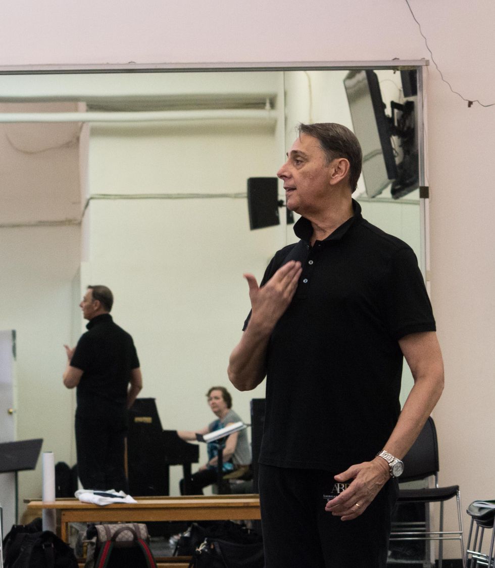 Raymond Lukens stands upright and holds his right hand to his breastbone while teaching a ballet class. He wears a black polo shirt and pants and stands in front of a dance studio mirror.