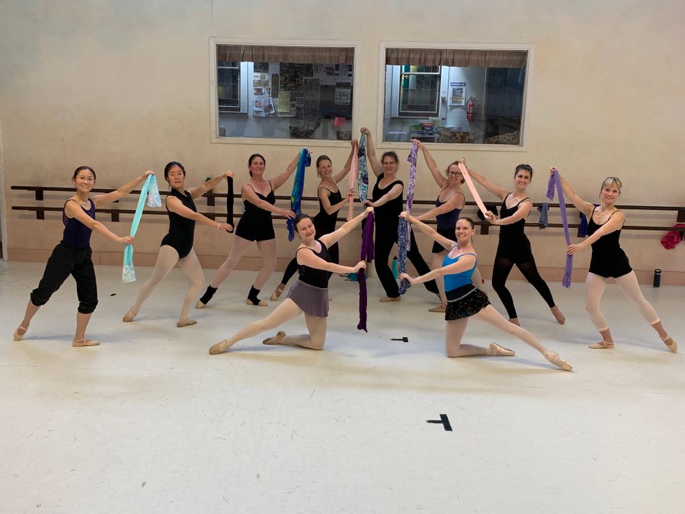 A group of 8 women lunge in tendu back with their arms in third position, holding a long scarf. Two other women kneel on the ground in a similar position with thier back leg and amrs. They all wear dark leotards, tights and some wear short skirts and warm-up pants.