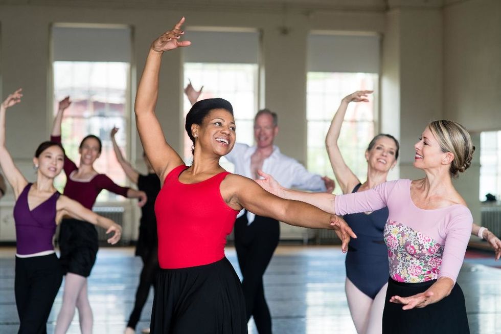 A woman in a red leotard and black ballet skirt smiles brightly in tendu croise while her teacher, wearing a floral pink leotard, stands to her left encouraging her.
