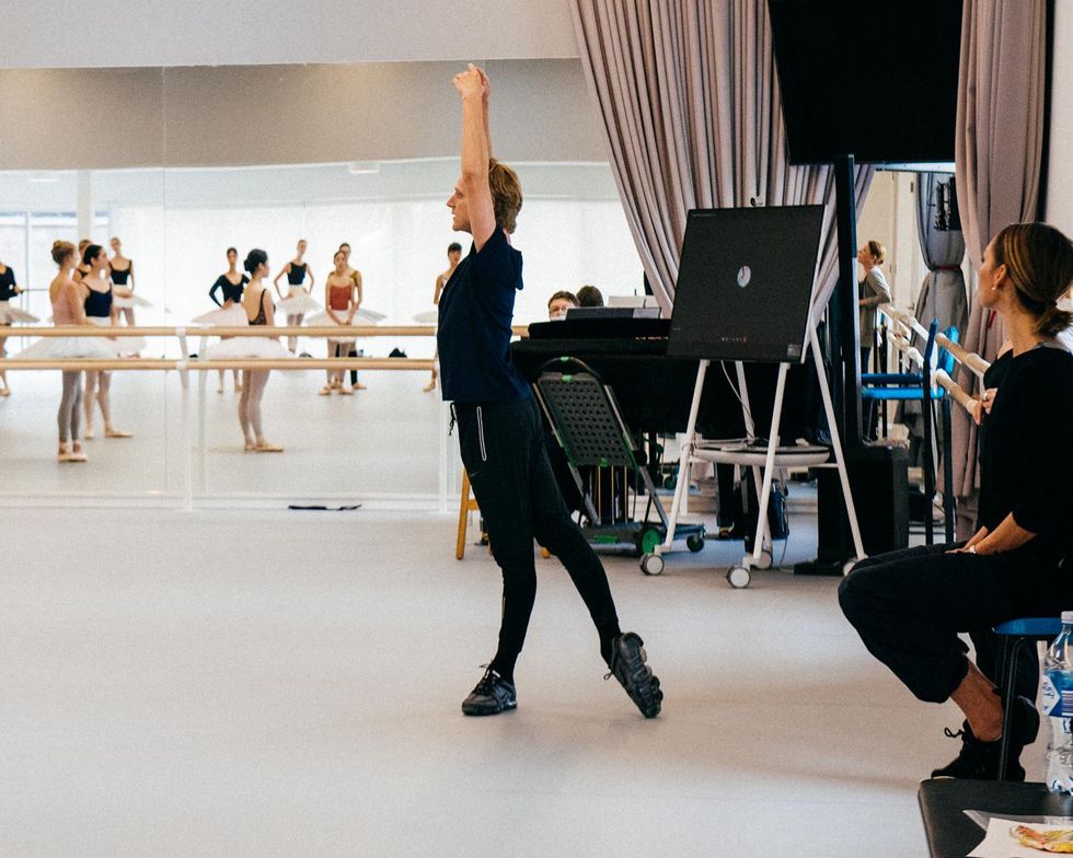 Wearing a black T-shirt, leggings and dance sneakers, David Hallberg stands in tendu derriere on his left leg with his arms in high fifth. A group of ballerinas in practice tutus watch.