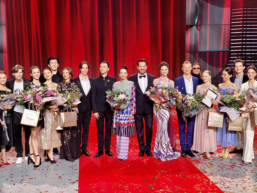 A staggered line of 17 men and women in dress clothes stand and smile for the camera in front of a red curtain. All of the women hold a bouquet of flowers, and confetti is scattered on the stage floor..